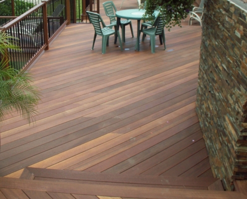 Batu-Deck-with-stacked-stone-wall a view looking down onto a multi-level batu deck. A stack stone wall with earth tones rises on the right side. The railing on the left has black, round, metal balusters.