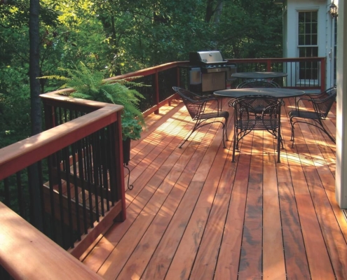 Tiger-Deck-In-the-Woods-with-metal-balusters A lush woodland scene closely surrounds a tropical hardwood tigerwood deck. The railing has black, round, metal balusters and a drink rail. Deck furniture and a BBQ sit on the deck.
